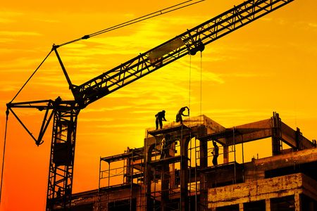 Worker on roof and crane on construction site silhouetted against orange sunset