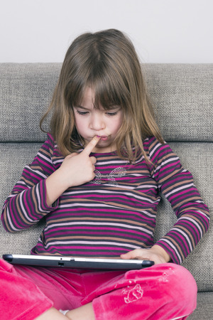 Little girl playing with a digital tablet at home on a sofaの写真素材