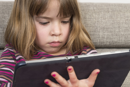 Little girl playing with a digital tablet at home on a sofaの写真素材