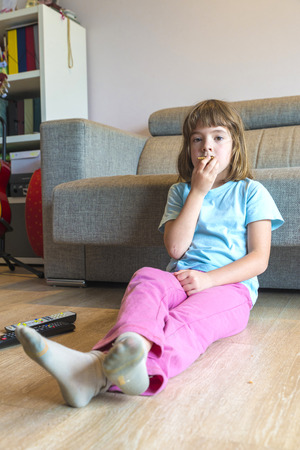 Little girl with TV remote control watching TV sitting on the parquet floor eating a cookieの写真素材