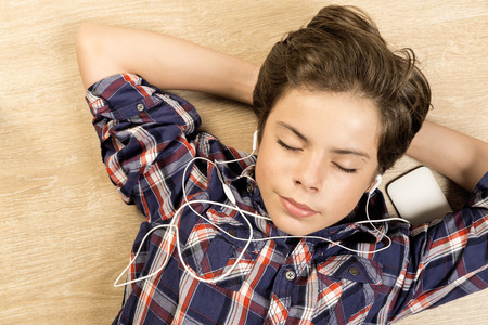 Portrait of a boy listening to music with headphones stretched on a wooden floor at home with arms behind his headの写真素材