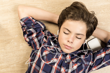 Portrait of a boy listening to music with headphones stretched on a wooden floor at home with arms behind his headの写真素材
