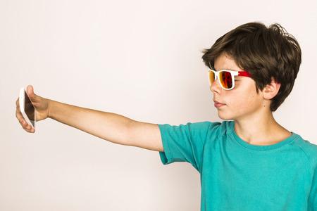 Happy young boy with sunglasses take a selfie with a smartphone at home isolated on white backgroundの写真素材