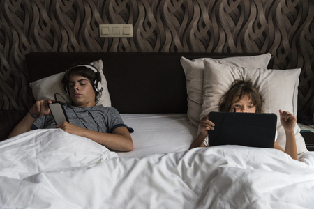 Boy and girl stretched out in bed relaxing and playing with smart phone and a digital tabletの写真素材