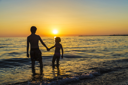 Silhouettes of a little girl and teenager bathing on a beach holding hands at sunset in summer in Sicily, Italyの写真素材