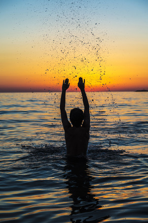 Silhouette of a teenager boy playing to throw water into the air on a beach at sunset in summer in Sicily, Italyの写真素材
