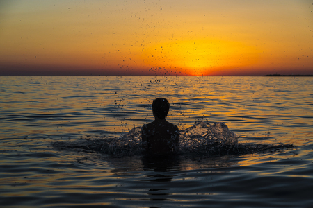 Silhouette of a teenager boy playing to throw water into the air on a beach at sunset in summer in Sicily, Italyの写真素材