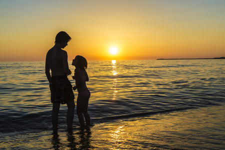 Silhouettes of a little girl and teenager bathing on a beach at sunset in summer in Sicily, Italyの写真素材