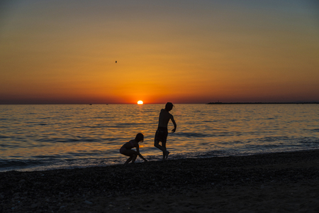 Silhouettes of a little girl and teenager playing throwing rocks at the sea on a beach at sunset in summer in Sicily, Italyの写真素材