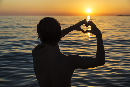 Silhouette of a teenager boy making the shape of a heart with his hands on a beach at sunset in summer in Sicily, Italyの写真素材