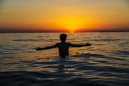 Silhouette of a teenager boy with arms outstretched on a beach at sunset in summer in Sicily, Italyの写真素材