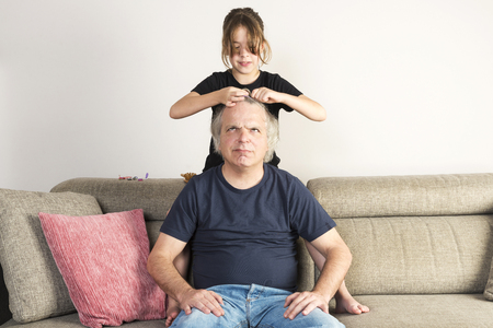 Little girl combing and making pigtails or braids to her dad on the couch at homeの写真素材