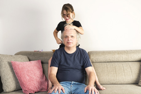 Little girl combing and making pigtails or braids to her dad on the couch at homeの写真素材