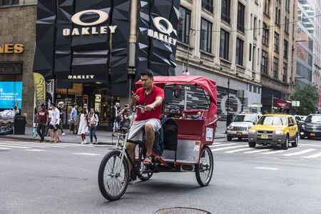 New York City, USA - July 28, 2018: Cyclist looking at the mobile phone while driving an electric tricycle on Fifth Avenue (5th Avenue) with people around and traffic in Midtown Manhattan, New York City, USAのeditorial素材