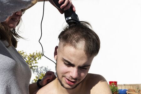 Mother cutting the hair of a teenager son with a hair clipper on the balcony of his home during the coronavirus quarantine in Barcelona, Catalonia, Spainの写真素材
