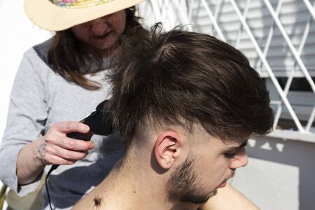 Mother cutting the hair of a teenager son with a hair clipper on the balcony of his home during the...