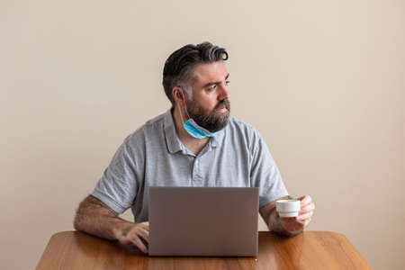 Mature Caucasian man drinks coffee, has coronavirus mask down, looks out the window and uses laptop