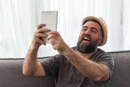man with beard, hipster, with tablet you take a photo selfie or have a video conference, video call. on the sofa, interior house.の写真素材