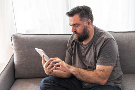white European man with a beard, sitting on a sofa, looks and presses a tablet, laptop.