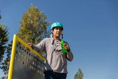 boy with roller skates, skateboard, is drinking water with green bottle in a public skate park. he has a protective helmetの写真素材