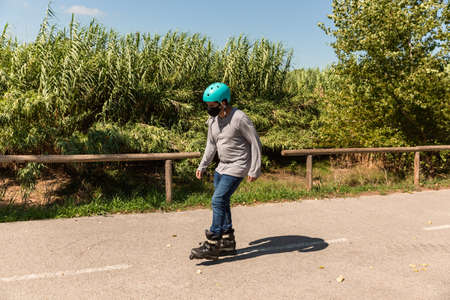 Young Caucasian Latino mix roller skater in a park, wearing a black mask for prevention of pandemic coronavirus and a cyan helmet, T-shirt and jeans.の写真素材