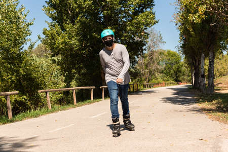 Young Caucasian Latino mix roller skater in a park, wearing a black mask for prevention of pandemic coronavirus and a cyan helmet, T-shirt and jeans.の写真素材