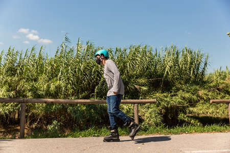 Young Caucasian Latino mix roller skater in a park, wearing a black mask for prevention of pandemic coronavirus and a cyan helmet, T-shirt and jeans.の写真素材