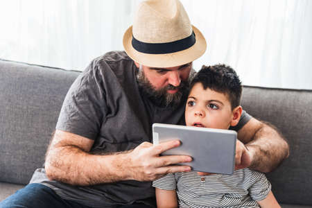 Caucasian bearded man and boy with a tablet on the sofa of a house. Casual dress, with hat.