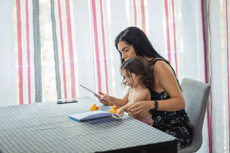 Hispanic mother and daughter use tablet sitting on a chair in front of a table in the dining room at home.の写真素材