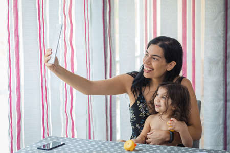 Hispanic mother and daughter use tablet sitting on a chair in front of a table in the dining room at home.の写真素材