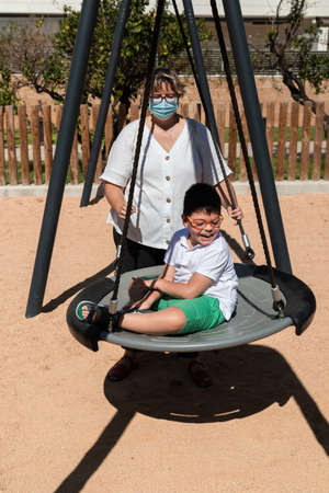 child with disability in swing with mother pushing, wears blue mask of pandemic coronavirus protection. playgroundの写真素材