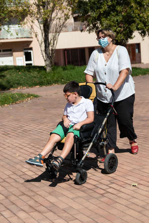 40-year-old Caucasian mother carries 5-year-old disabled boy in wheelchair along city sidewalk during walk. She wears mask for prevention of coronavirus pandemic.の写真素材