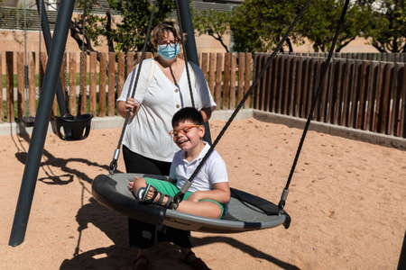 child with disability in swing with mother pushing, wears blue mask of pandemic coronavirus protection. playgroundの写真素材