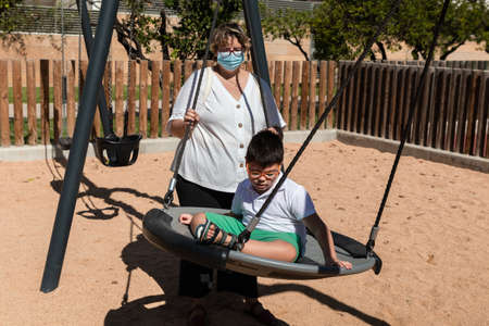 child with disability in swing with mother pushing, wears blue mask of pandemic coronavirus protection. playgroundの写真素材