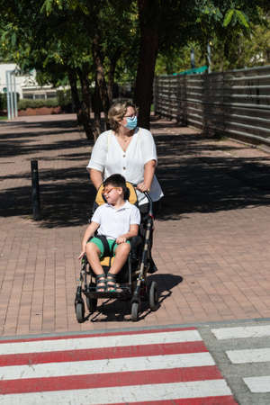 40-year-old Caucasian mother carries 5-year-old disabled boy in wheelchair along city sidewalk during walk. She wears mask for prevention of coronavirus pandemic.の写真素材