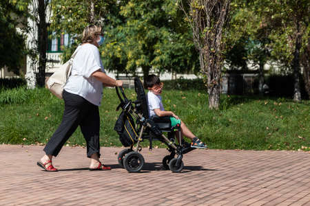 40-year-old Caucasian mother carries 5-year-old disabled boy in wheelchair along city sidewalk during walk. She wears mask for prevention of coronavirus pandemic.の写真素材