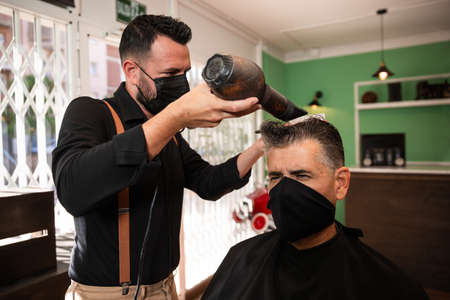 white caucasian barber combs and blow-drys another man's hair in a barbershop during the coronavirus pandemic. they go retro hipster style and both wear black masks.の写真素材