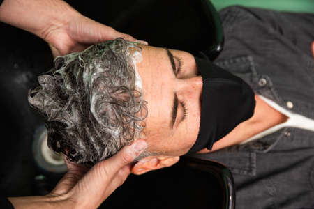 White Caucasian man with black mask of protection pandemic coronavirus is lying down while some barber's hands wash his hair with water and soap in hairdresser's, barber shop.の写真素材