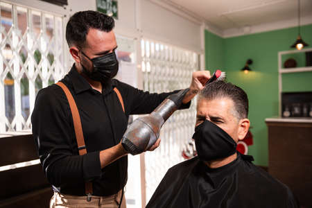 white caucasian barber combs and blow-drys another man's hair in a barbershop during the coronavirus pandemic. they go retro hipster style and both wear black masks.の写真素材