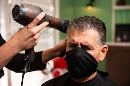 white caucasian barber combs and blow-drys another man's hair in a barbershop during the coronavirus pandemic. they go retro hipster style and both wear black masks.の写真素材