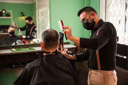 white caucasian barber combs and blow-drys another man's hair in a barbershop during the coronavirus pandemic. they go retro hipster style and both wear black masks.の写真素材