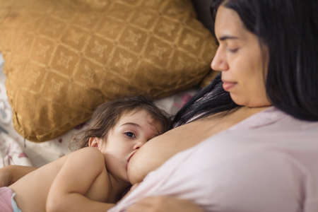 Latino mother breastfeeds her baby girl in the double bed in a pink gown.