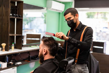 Hispanic barber combs and dries the hair of the head to Caucasian client with beard in a green,...