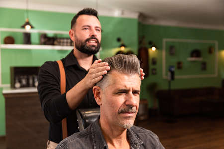 barber with shirt and suspenders comb with brush and hands to man with gray hair and goatee in a...