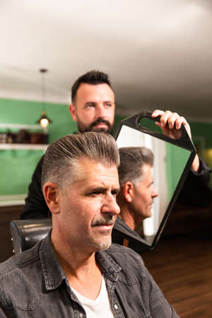 Bearded barber shows haircut and hairstyle to his client with a goatee with a mirror in the barbershop, both Caucasian.の写真素材