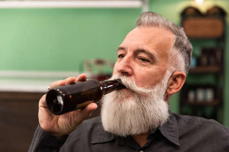 Older Caucasian bearded man, bearded model, in a retro hipster bar drinks a bottle of beer, photo portrait.の写真素材