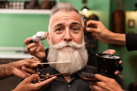 concept of barbershop, an older man with a white beard is shaved, combed, hair cut by several hands of hairdressers and barbers. multiethnic people. barbering toolsの写真素材