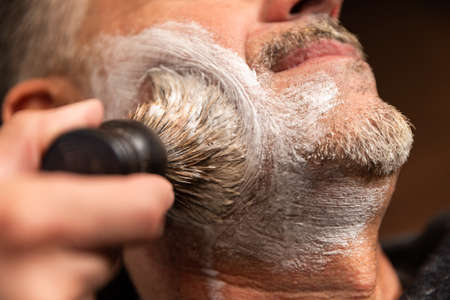 barbershop, barber's hand puts shaving foam with a razor blade on a client's cheek, both Caucasian. the client is lying on the chair and wearing a black gown.の写真素材