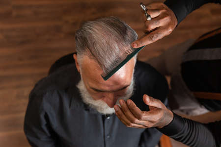 African-American barber with tattooed hands combs the head hair of a gray-haired, bearded Caucasian man, seen from above.の写真素材
