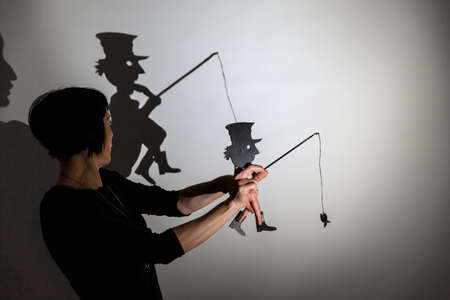 play shadow projected on a white screen. the person's hands shape a poor man, a homeless man, with a hat and a rod fishing with a dead fish skeletonの写真素材